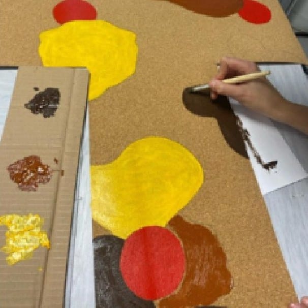 Hands painting yellow and red shapes onto a cork panel