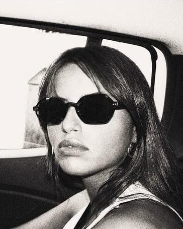 Black and white photo of Maria Loureiro in sunglasses, looking back over her shoulder from the seat of a car